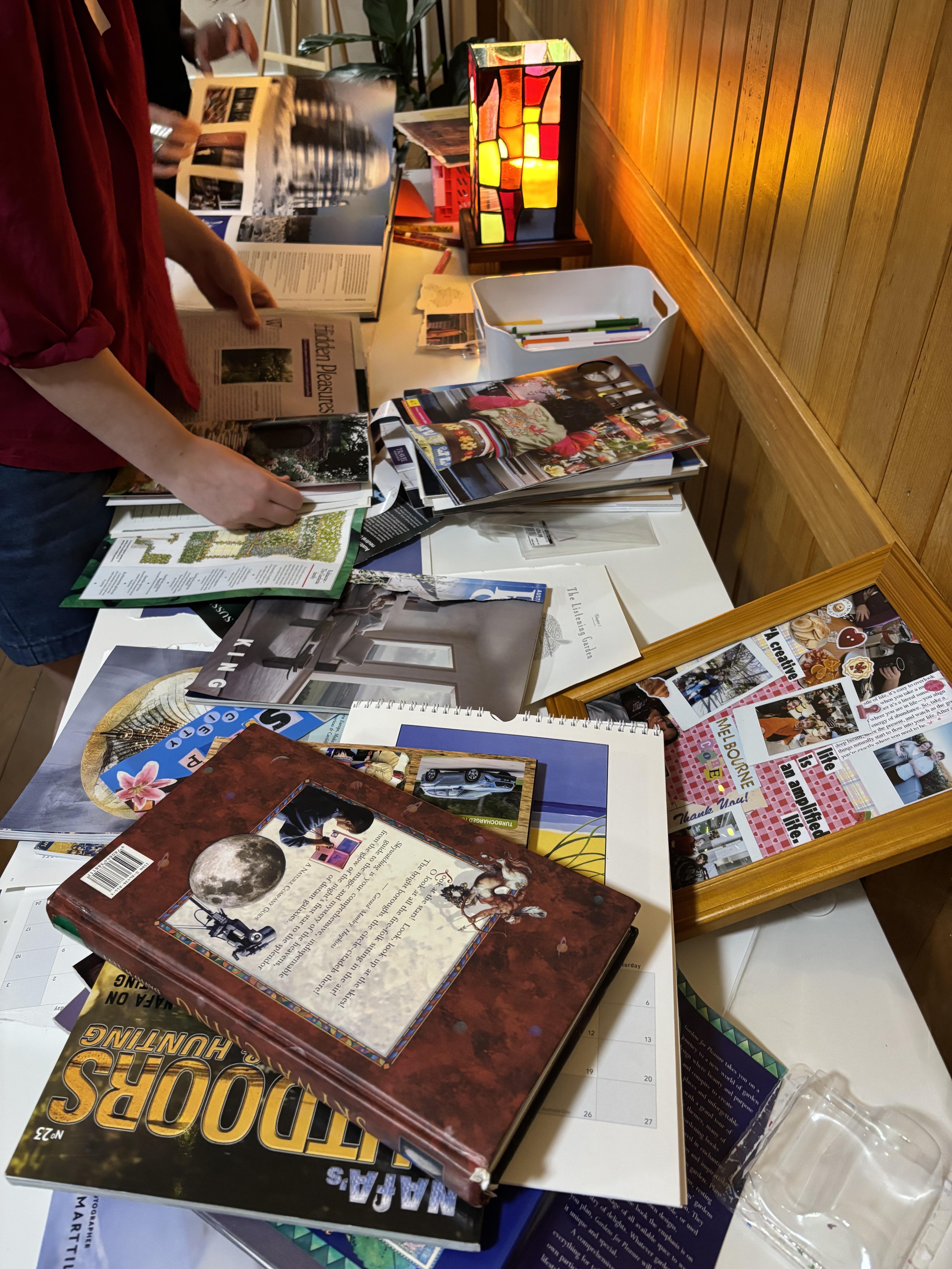 A variety of old books, calendars, pens are stacked on a table with a bright coloured lamp glowing in the corner