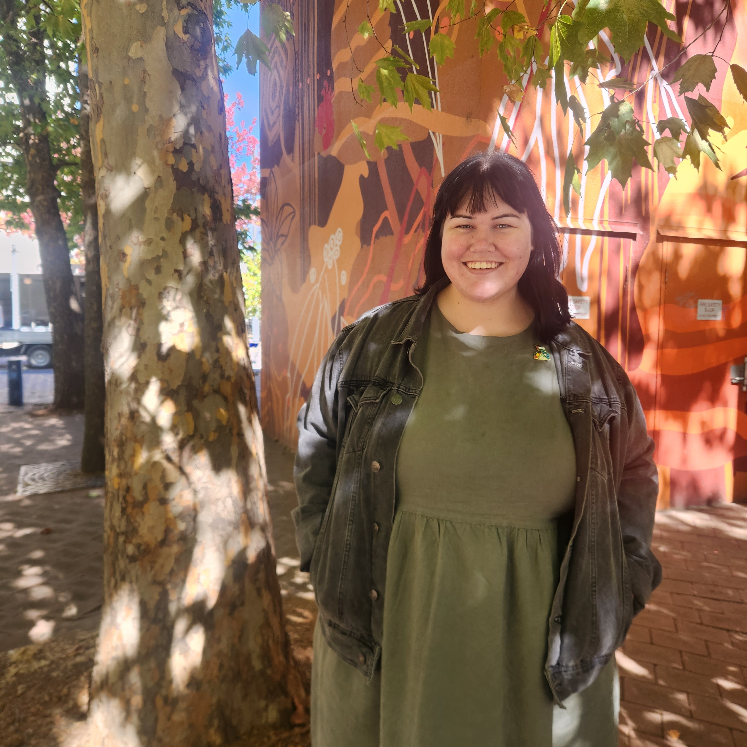 A young woman wearing a green dress and black jacket stands near a tree.