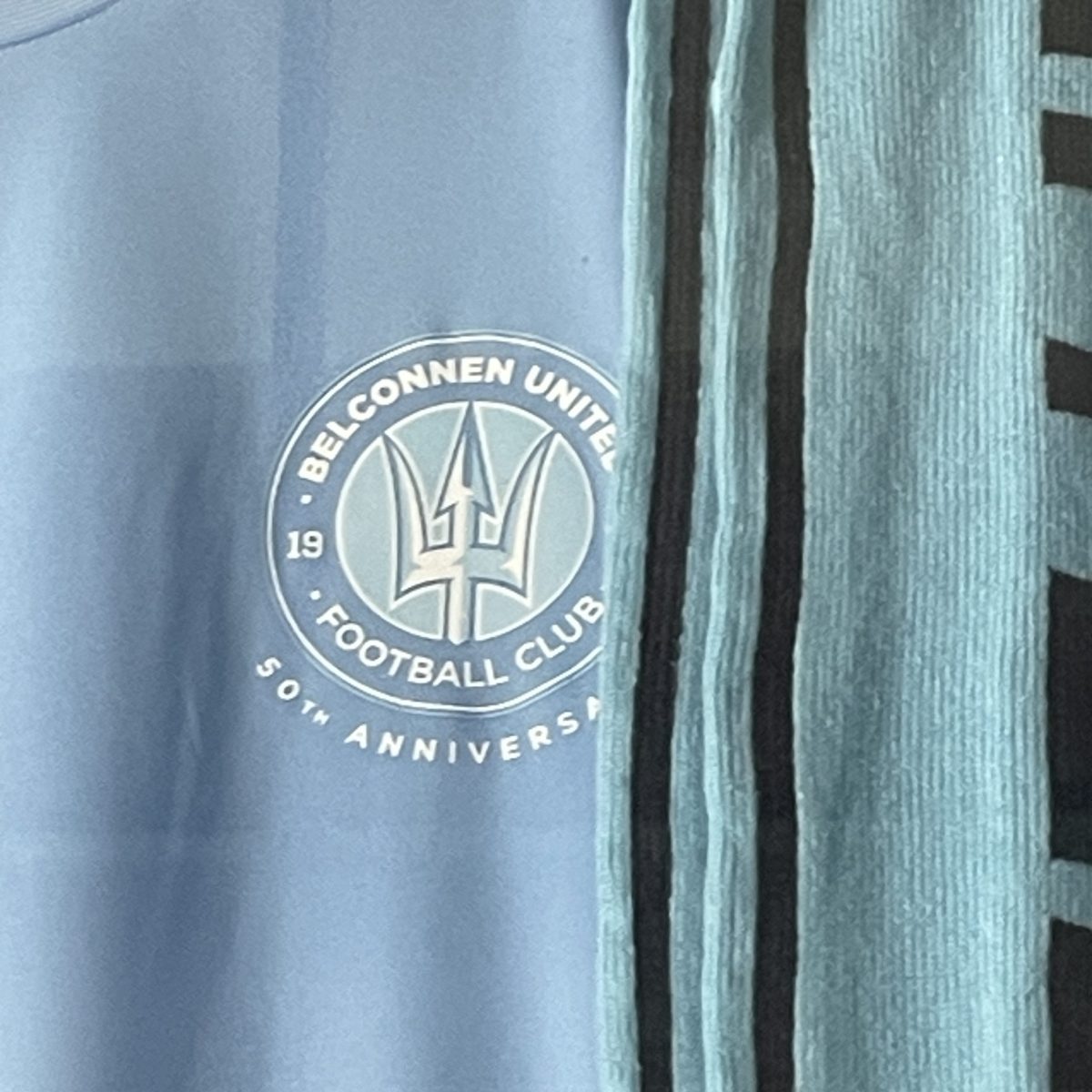 A light blue football shirt with a circular logo featuring a trident in the middle surrounded by the words Belconnen United Football club. Light blue scarf next to shirt