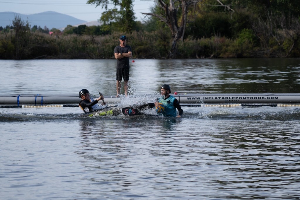 Two people clash into each other in kayaks while a man is standing behind them with his arms cross standing on a pontoon