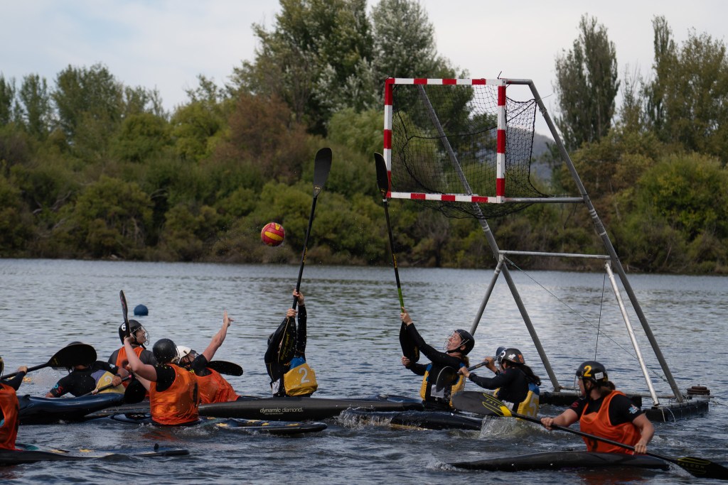Ball in air heading towards a set of Canoe polo goals but is being blocked by a paddle being held up by a person in a kayak with many other people around them in kayaks
