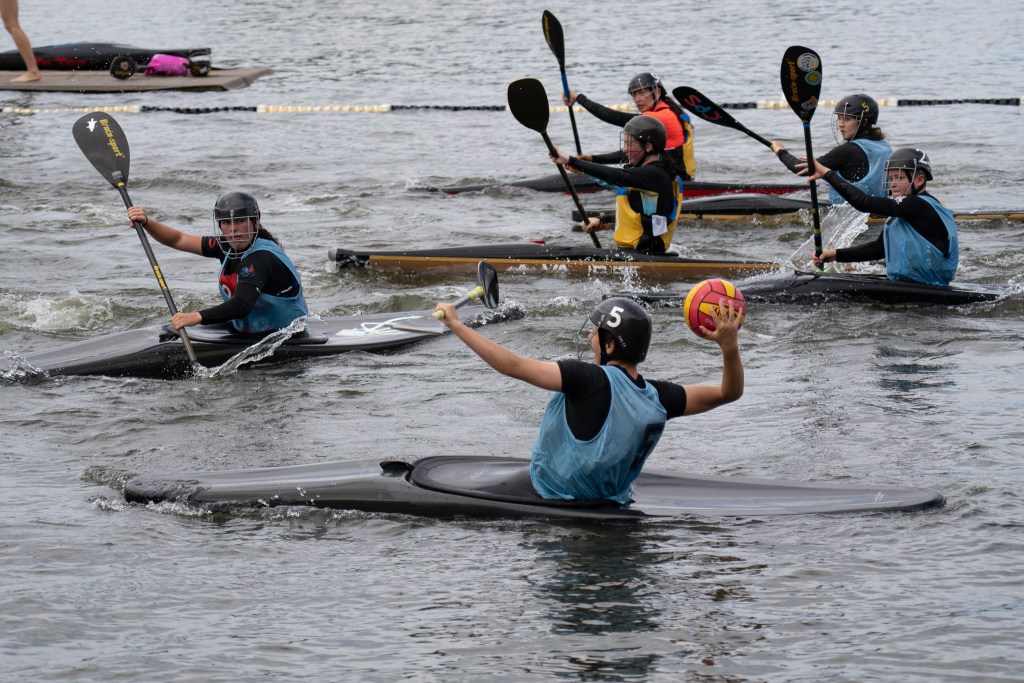 Person in blue shirt and wearing a helmet is throwing a ball while in a kayak surrounded by people in kayaks