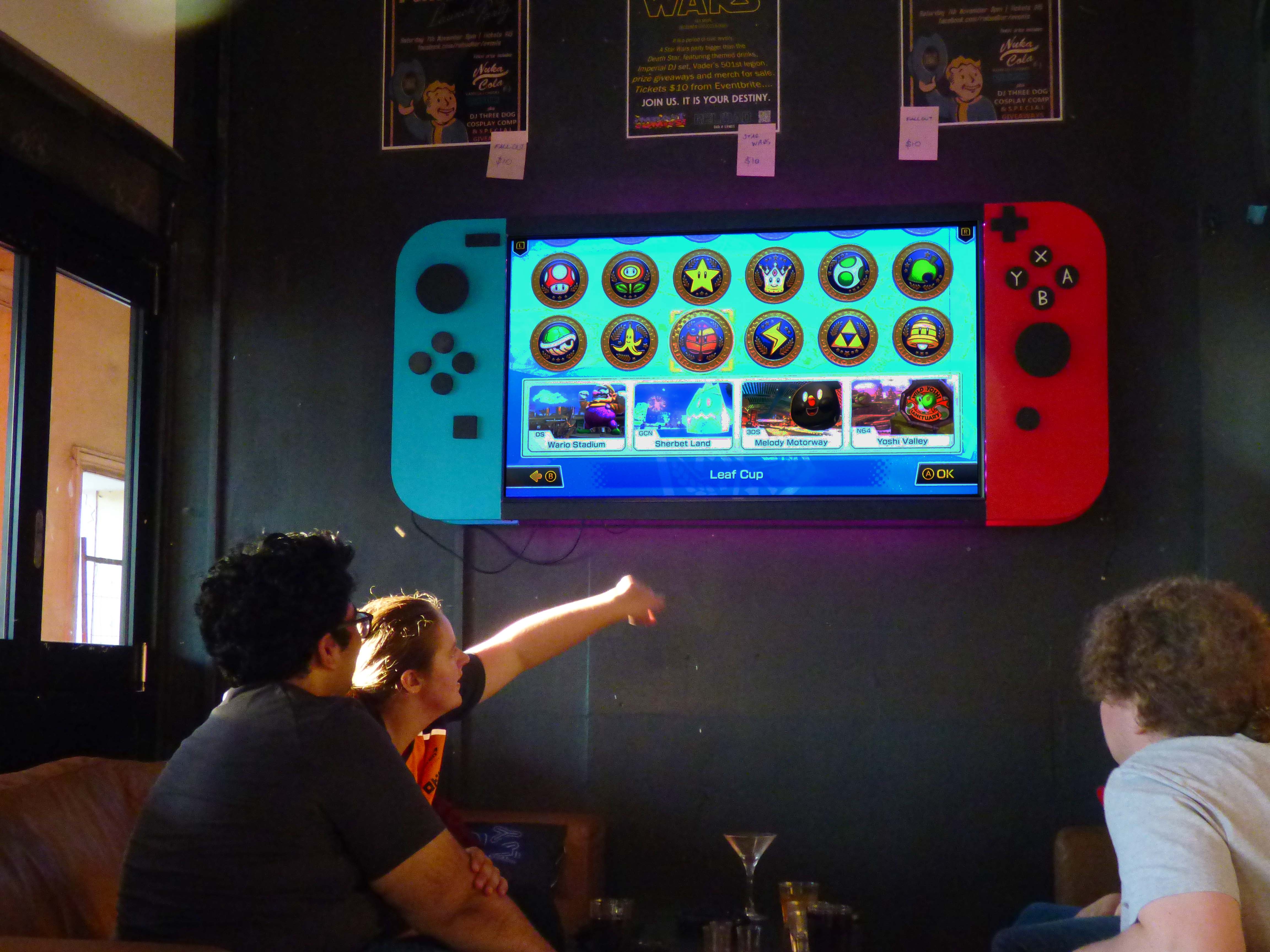 Three people playing Mario cart on a wall mounted television that looks like a giant Nintendo switch. 