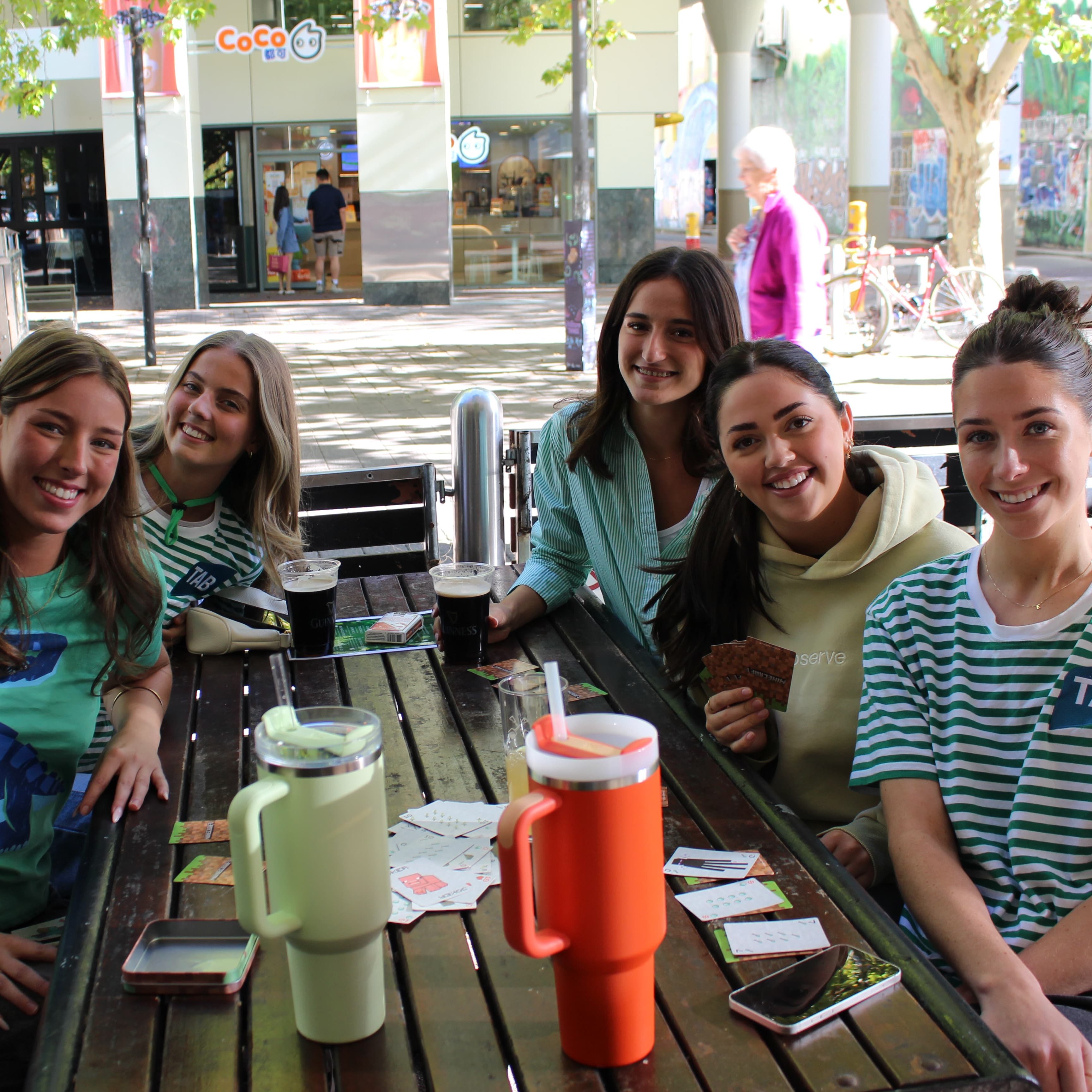 A group of girls sit around a table outside with a card game set out.