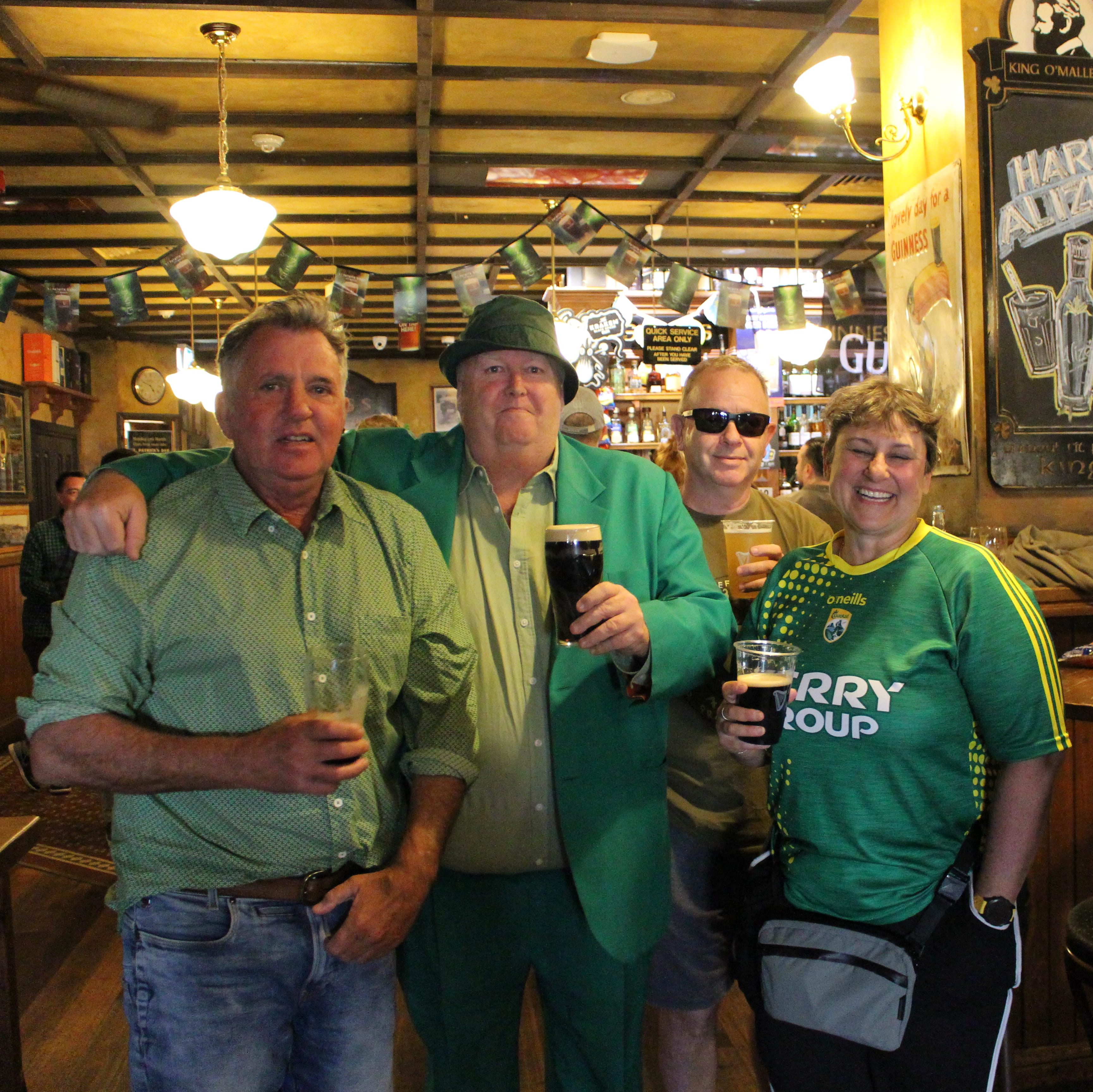 A group of four older patrons, wearing green attire, pose together with their pints of Guiness.