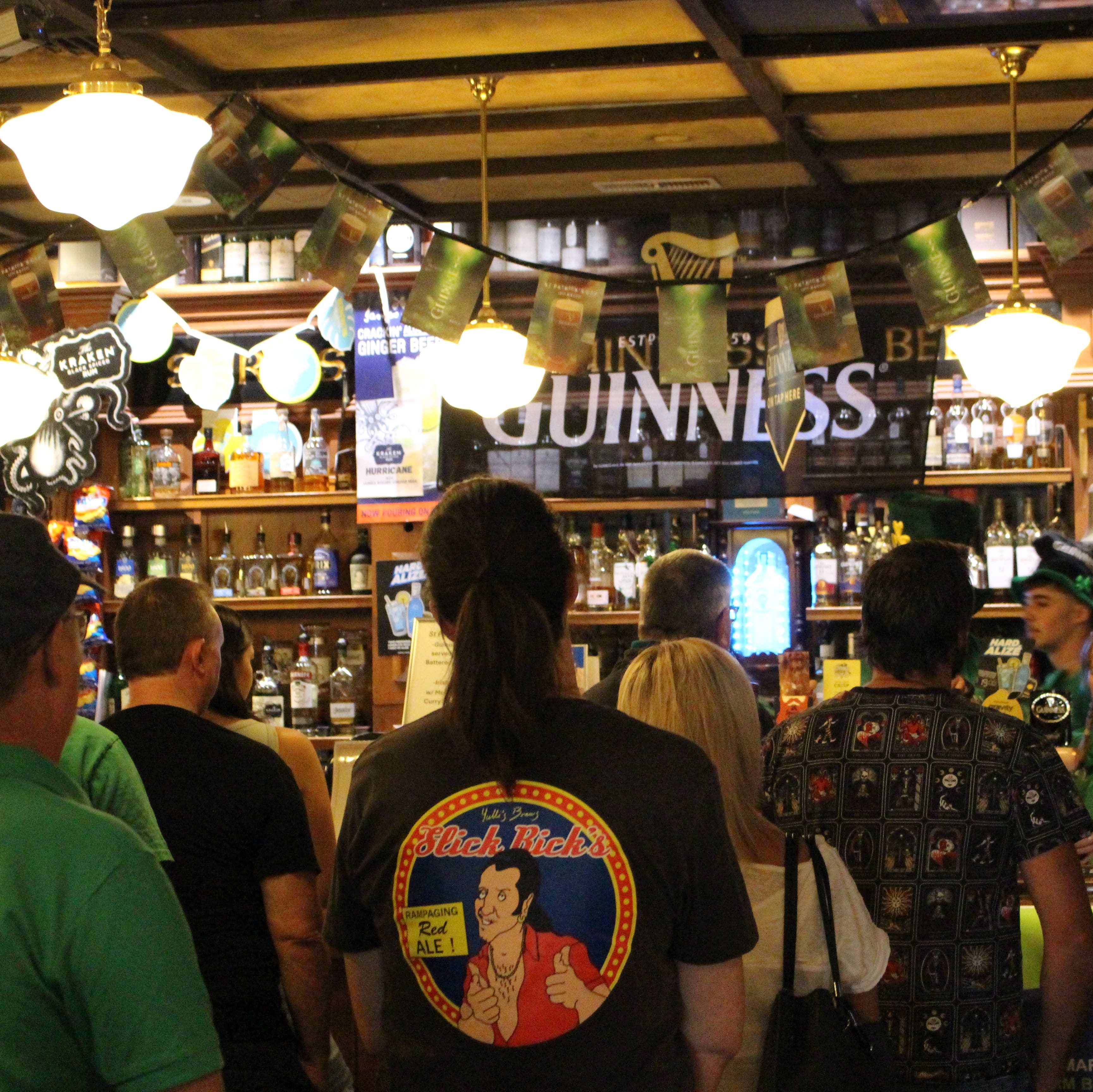 A crowd of patrons line up in front of the bar to order their next drink. A big Guinness flag hangs in front of the liquors.