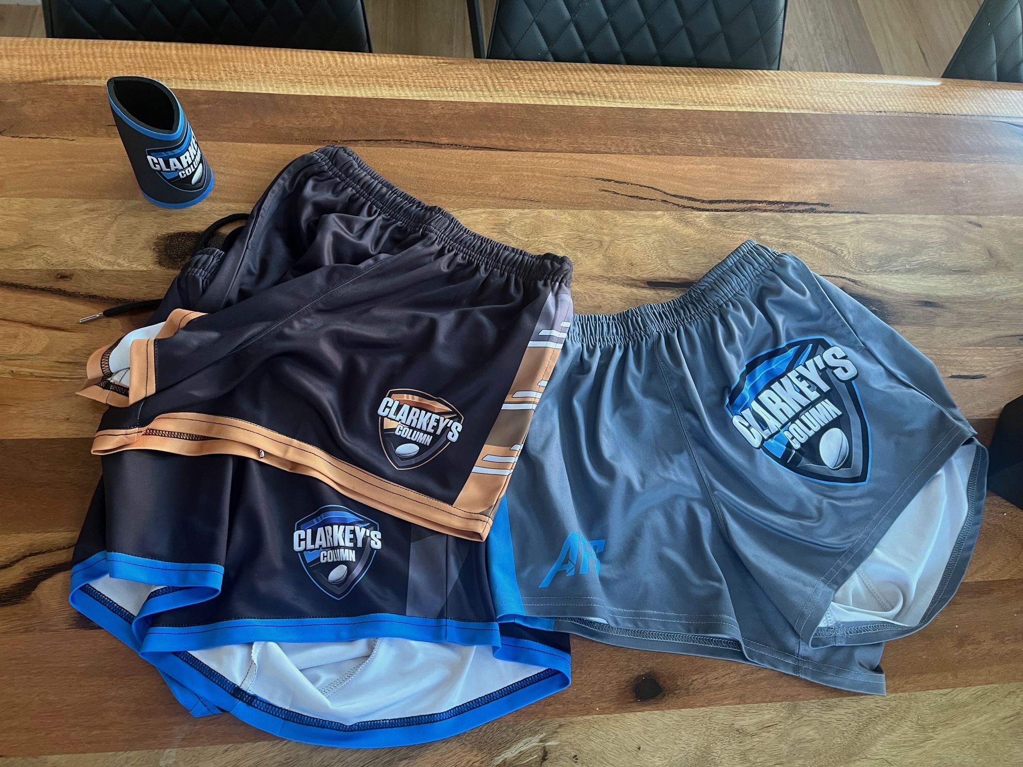 Three pairs of shorts and a can cooler all laid out on a wooden table, each have Clarkey's logo on them. 