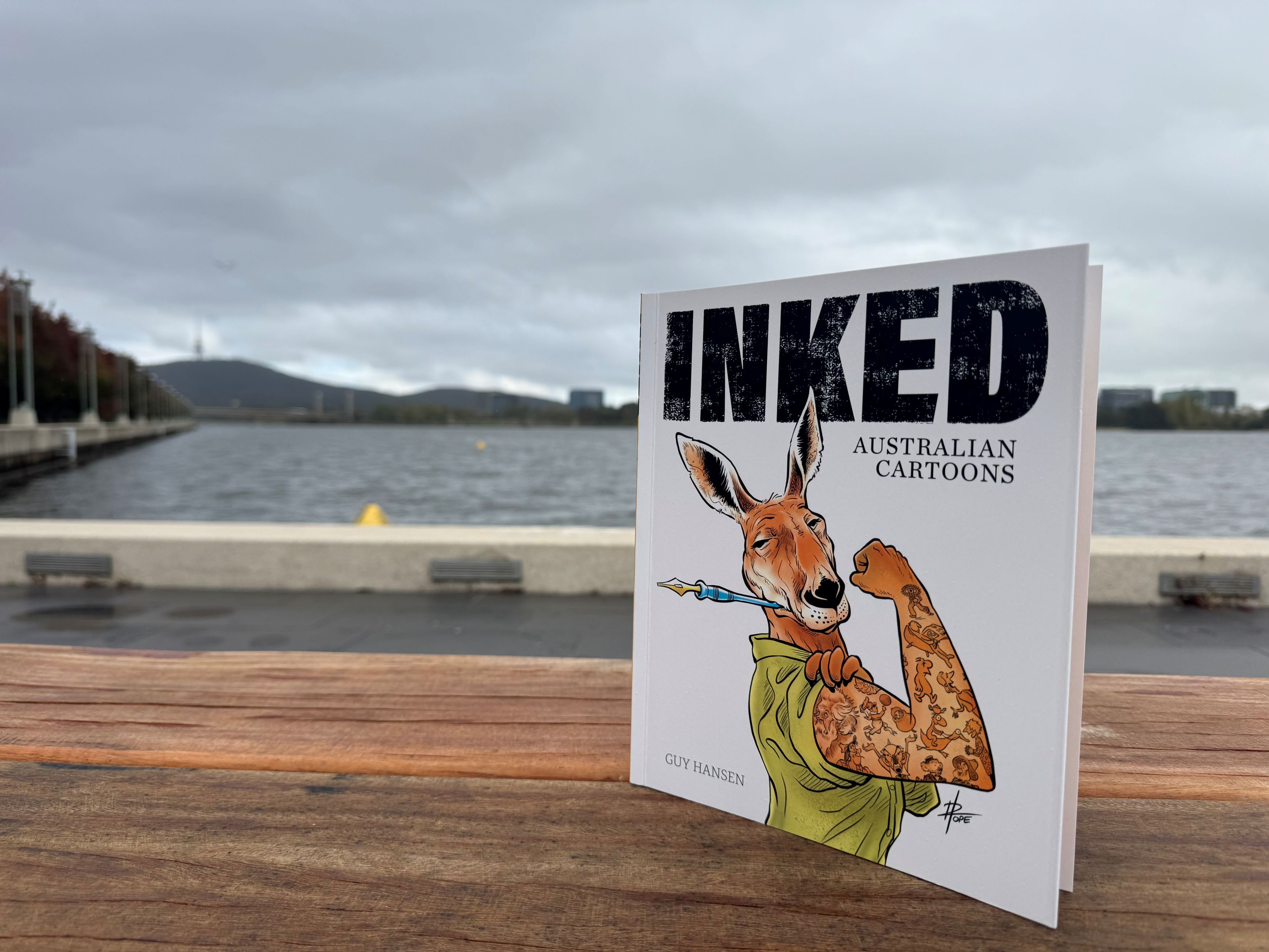 A copy of Guy Hansen's book Inked in the foreground with Lake Burley Griffin in the background