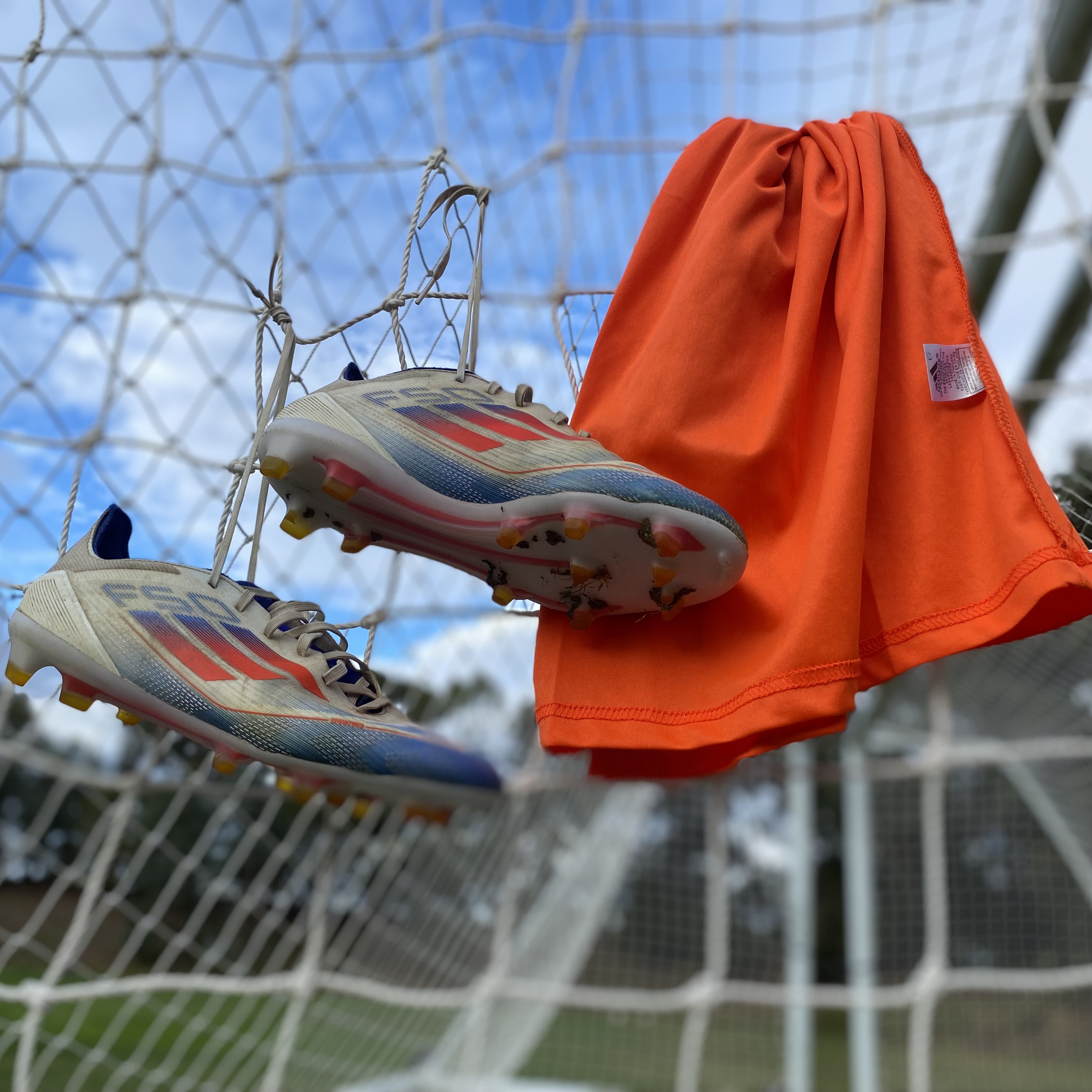 Boots hanging from a net with an orange jersey in the background, with blurred treelines further back
