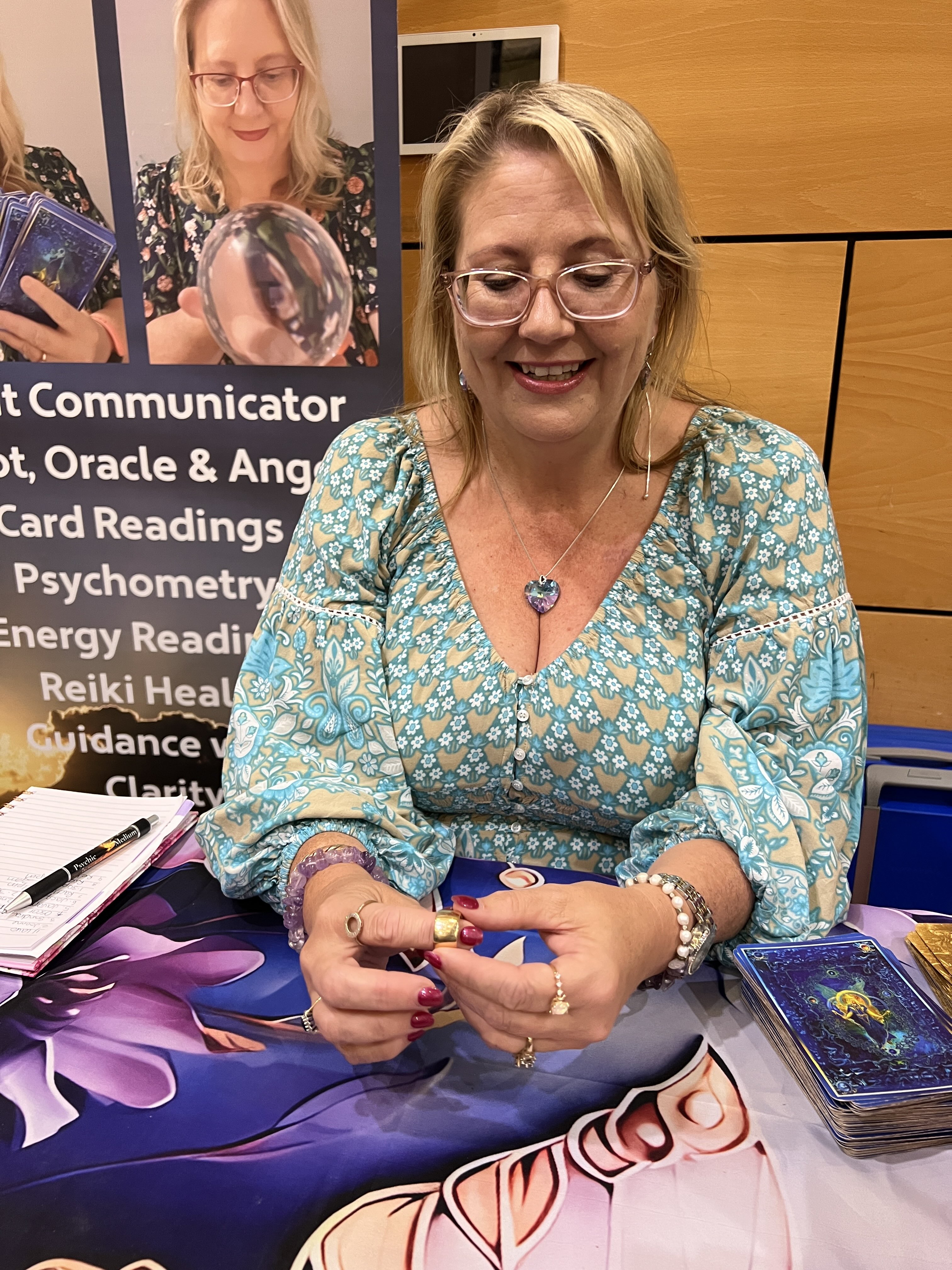 Blonde woman wearing glasses and blue shirt smiling and holding gold ring