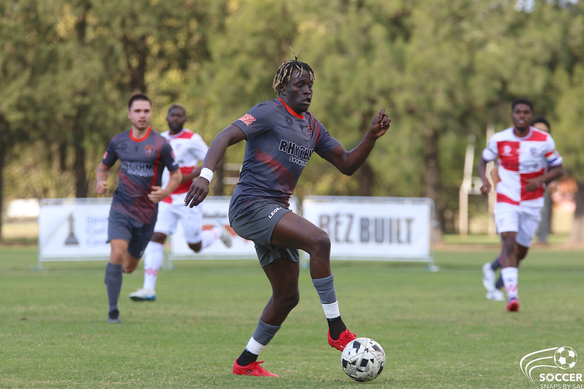 Akio stretches out his left foot to control the ball with red boots on a football field, with teammates and opposition players in the background.