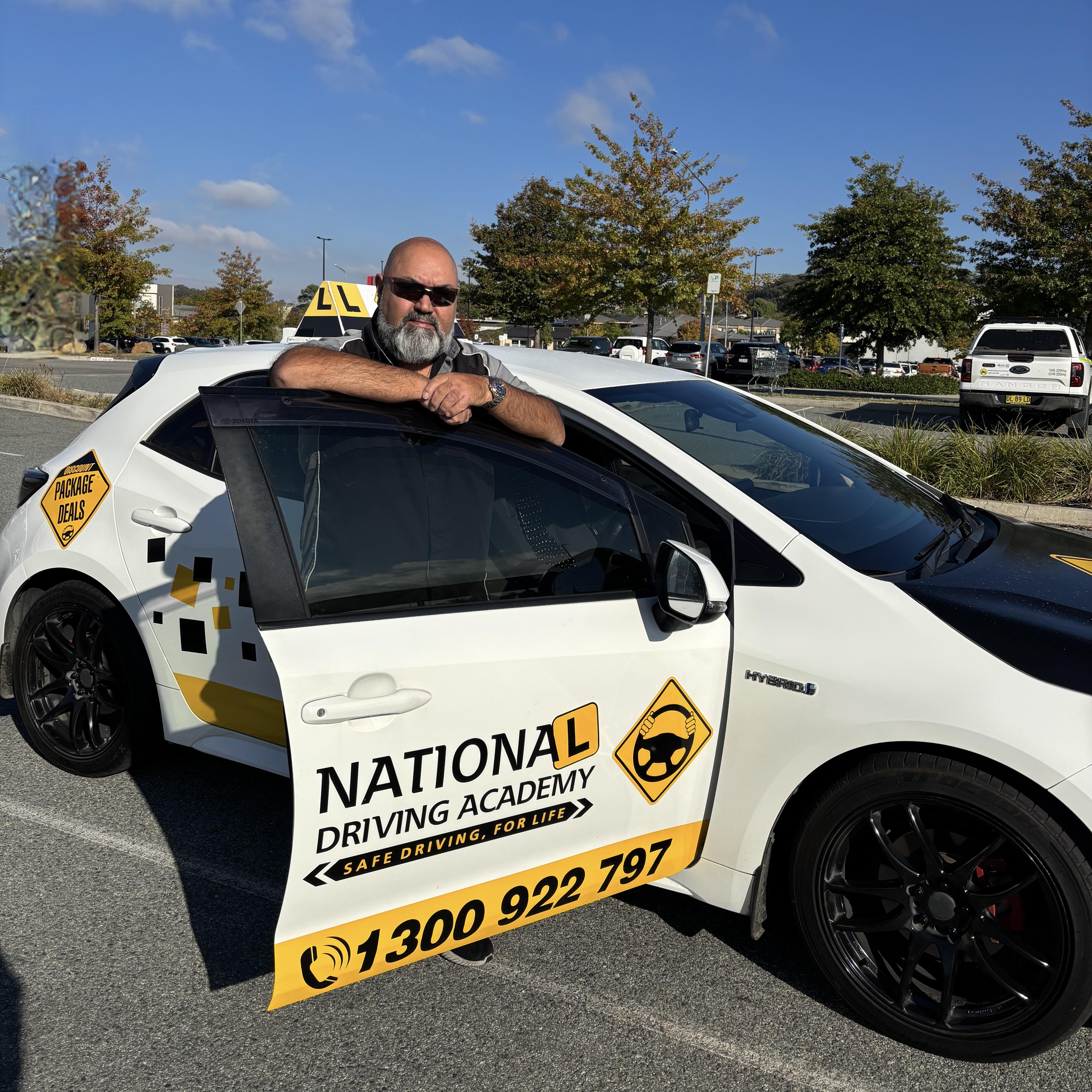 A male driving instructor (Babar) with sunglasses leans on an open door that reads NATIONAL DRIVING ACADEMY