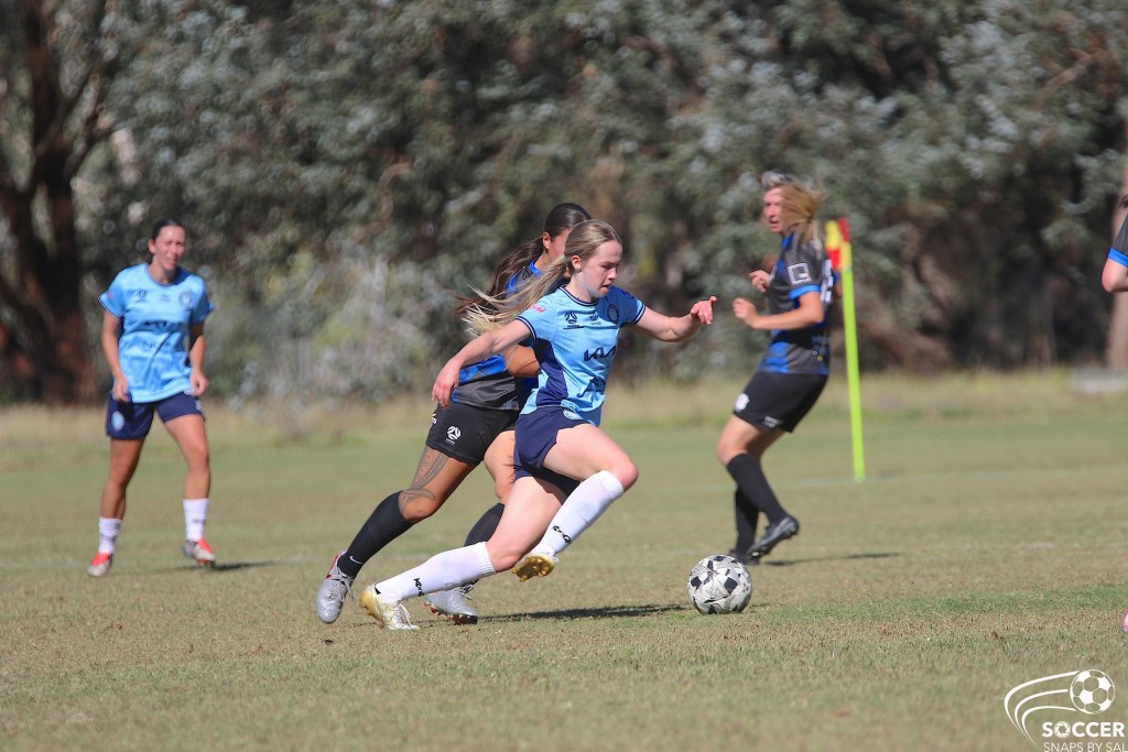 A female soccer player in a light blue jersey dribbles the ball forward on a grassy field, with defenders in dark jerseys pursuing her.