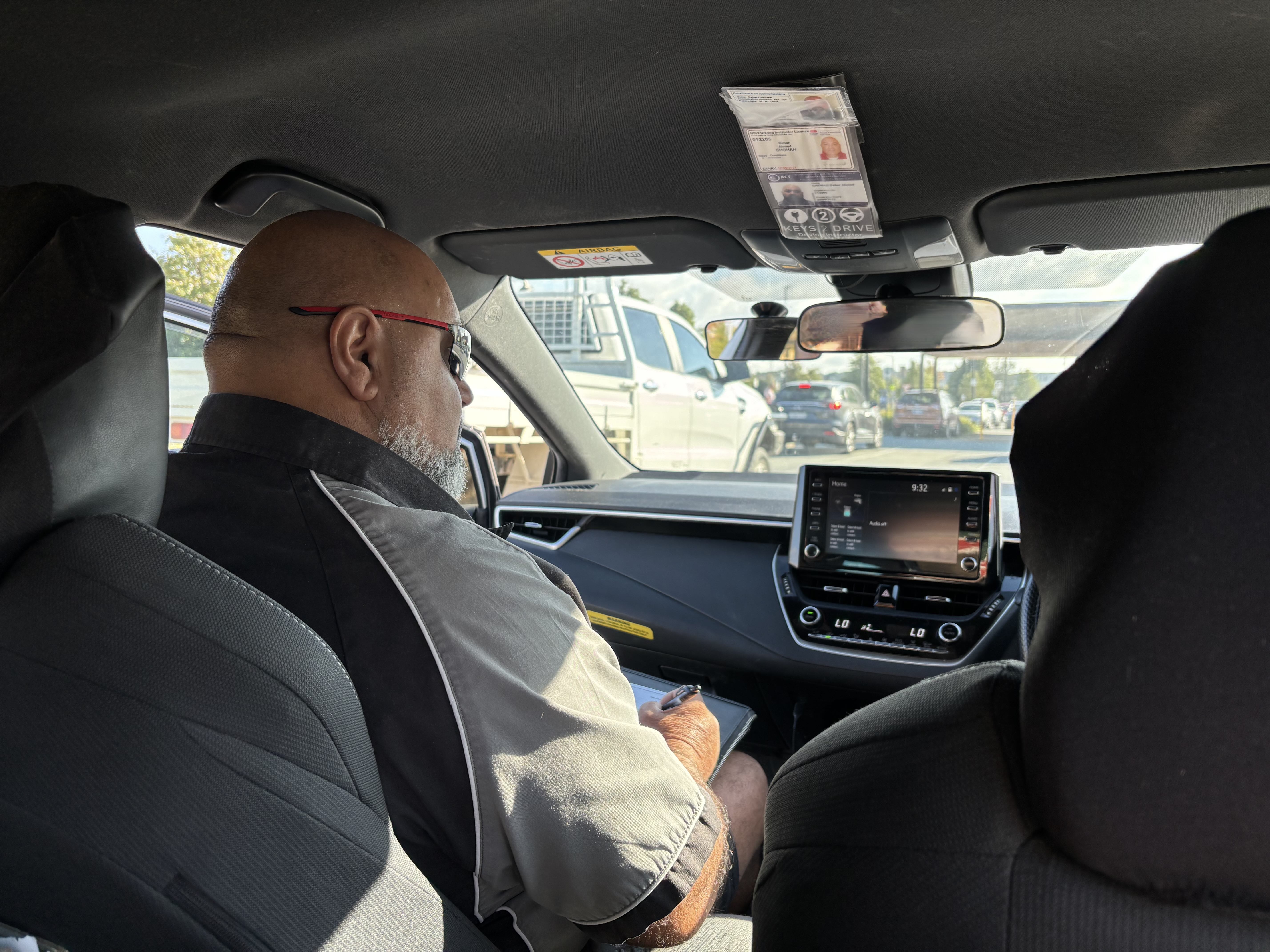 A male driving instructor (Babar) sits in the passenger side of the car looking out the window.