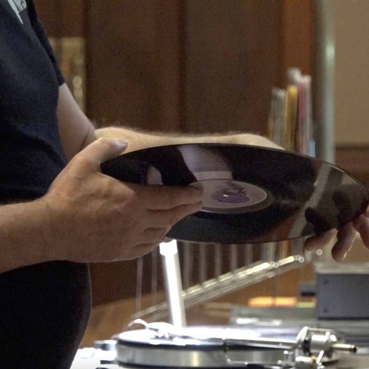 A man's hands hold a black vinyl. Behind him is a record player.
