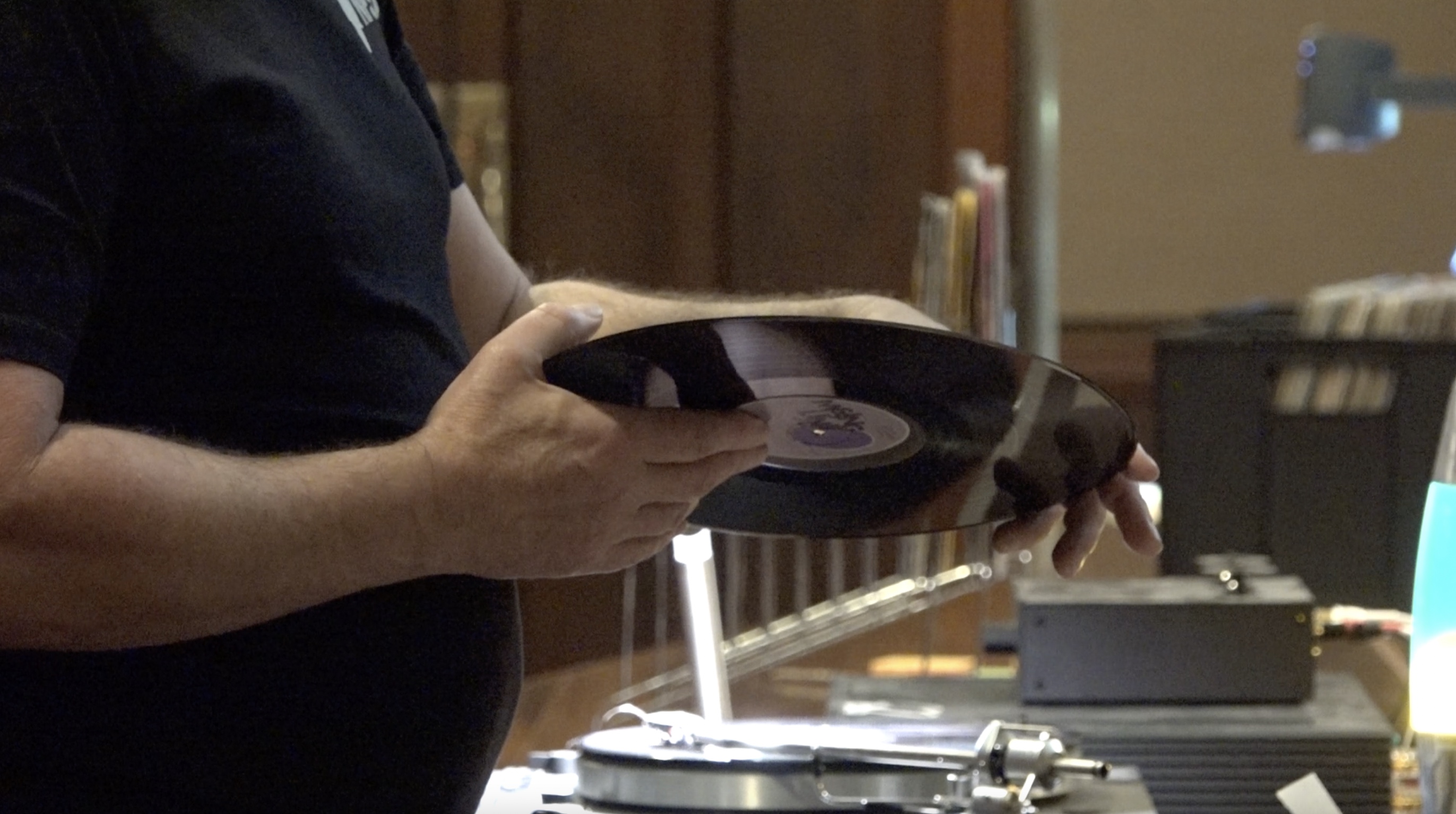 A man holds a black shiny vinyl. In the background are blurred crates with more records in them
