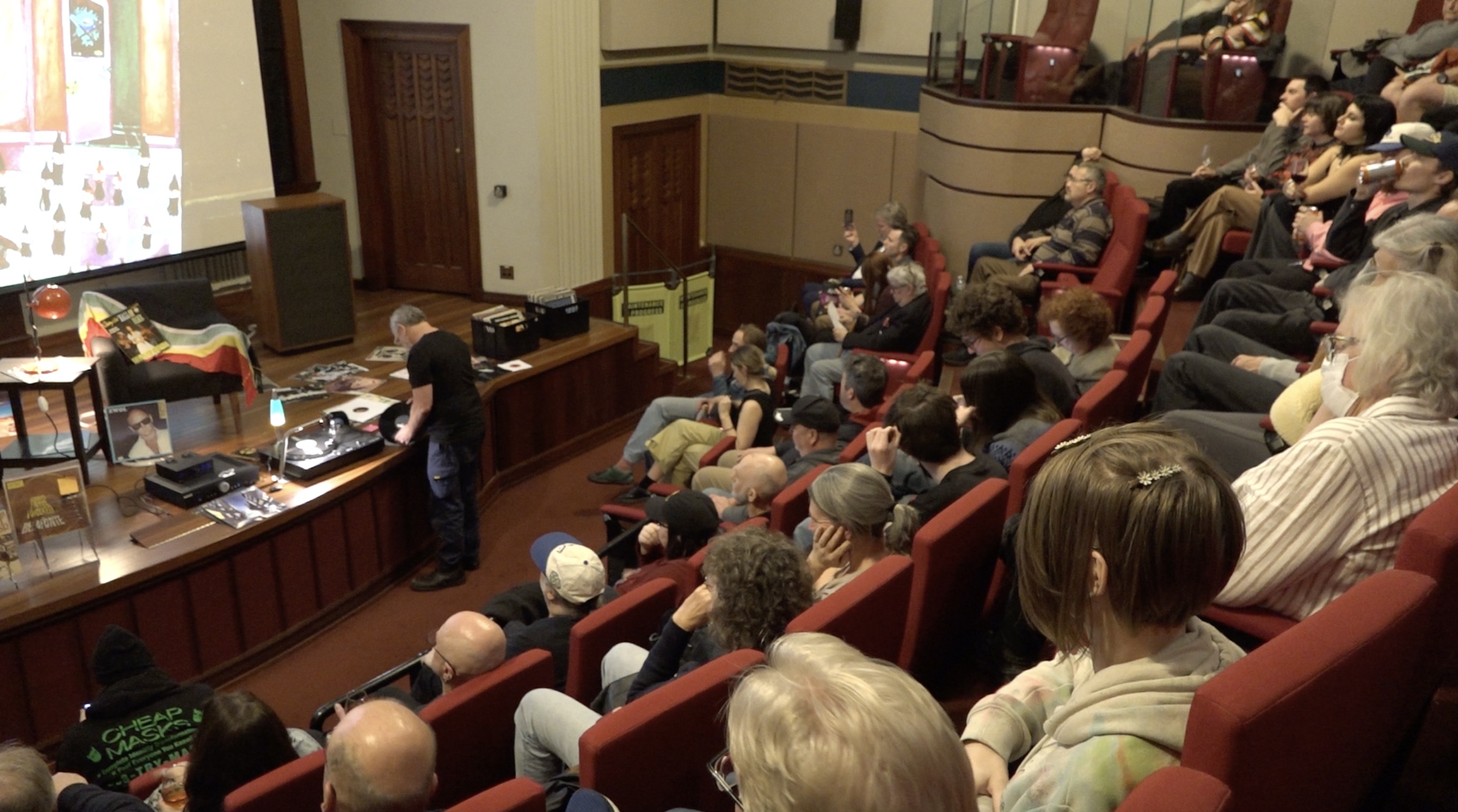 The camera looks down on the crowd from above and behind. People's attention is focused on the stage where a record player is set up and records are on display.