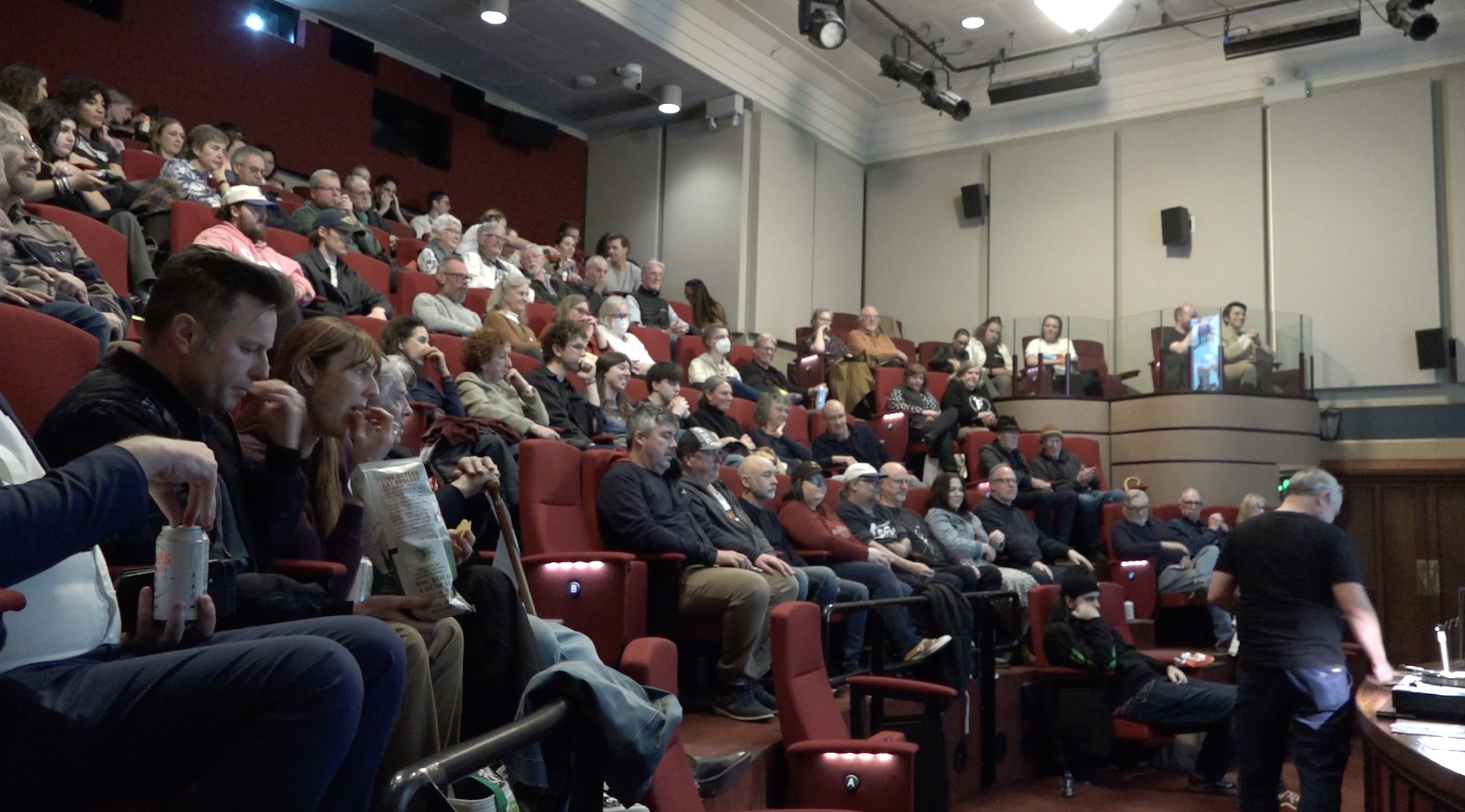 A crowd of people aged from their 20s to 70s sits on red plush tiered seats. They all look towards the front of the room