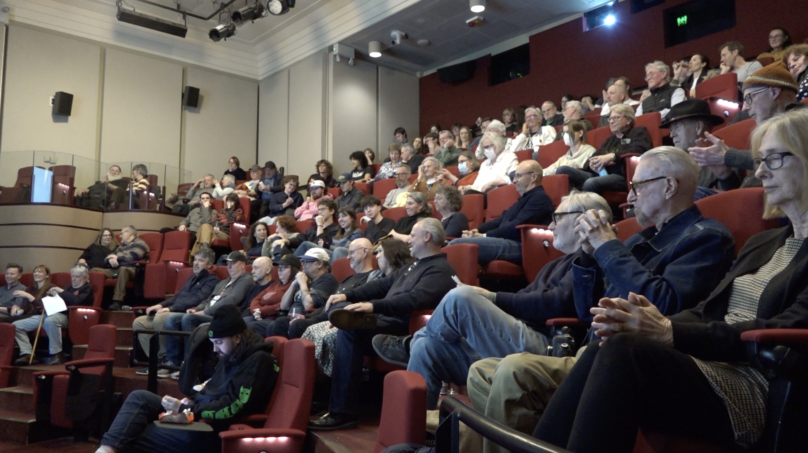 A crowd sit on red plush tiered seats. Some of them are clapping.