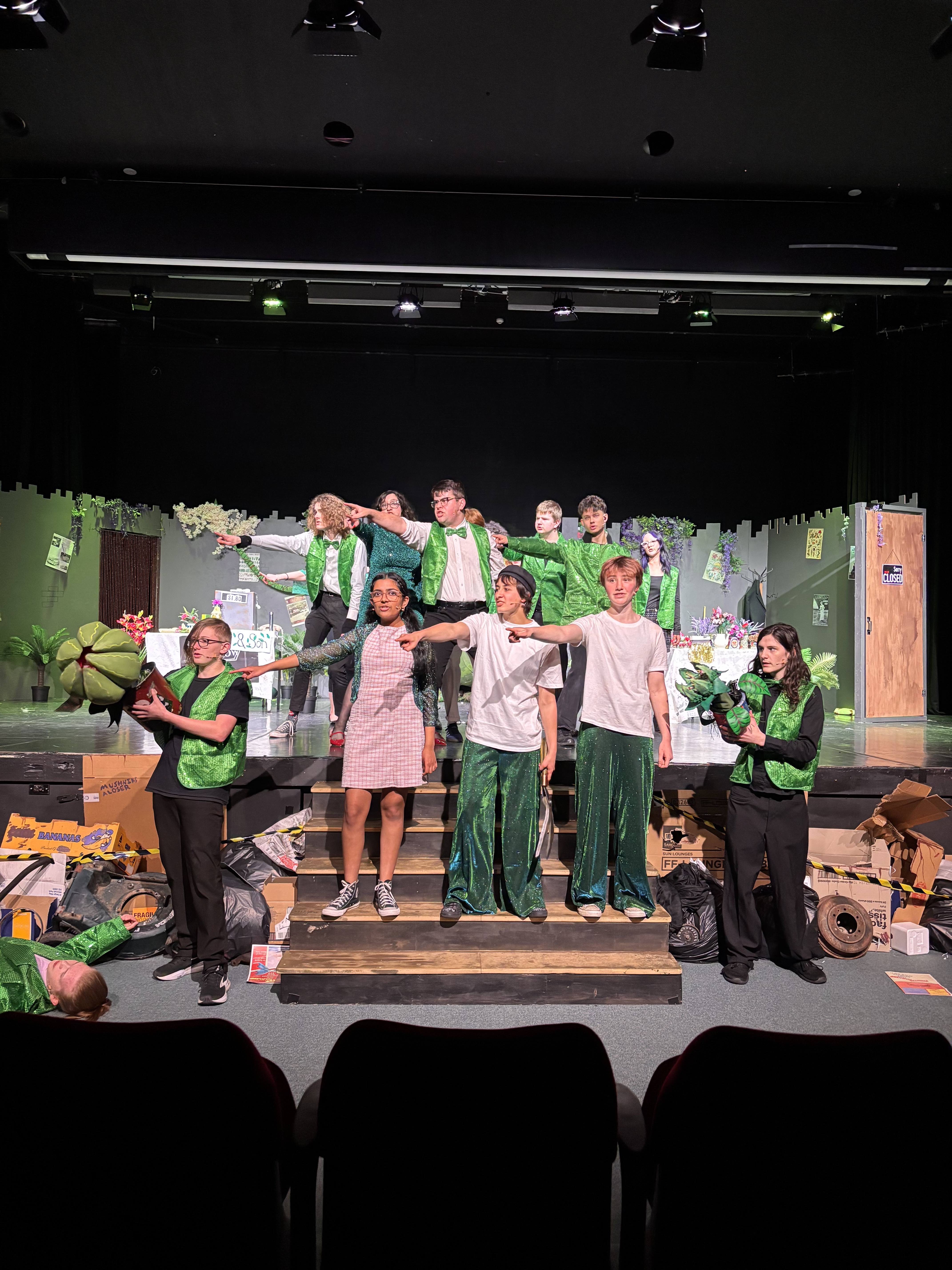 A group of young men and women stand on stage wearing various green sequinned accessories. They are mid-dance.