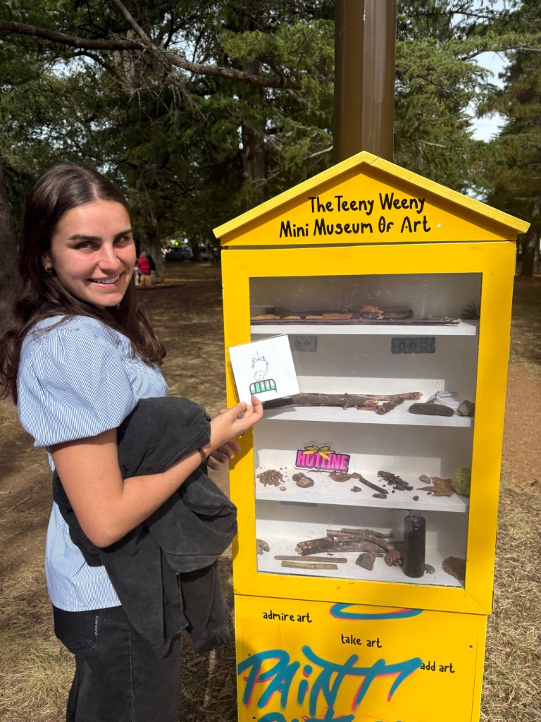 Photograph of a girl smiling next to an outdoor bookshelf that reads "the teeny weeny mini museum of art"