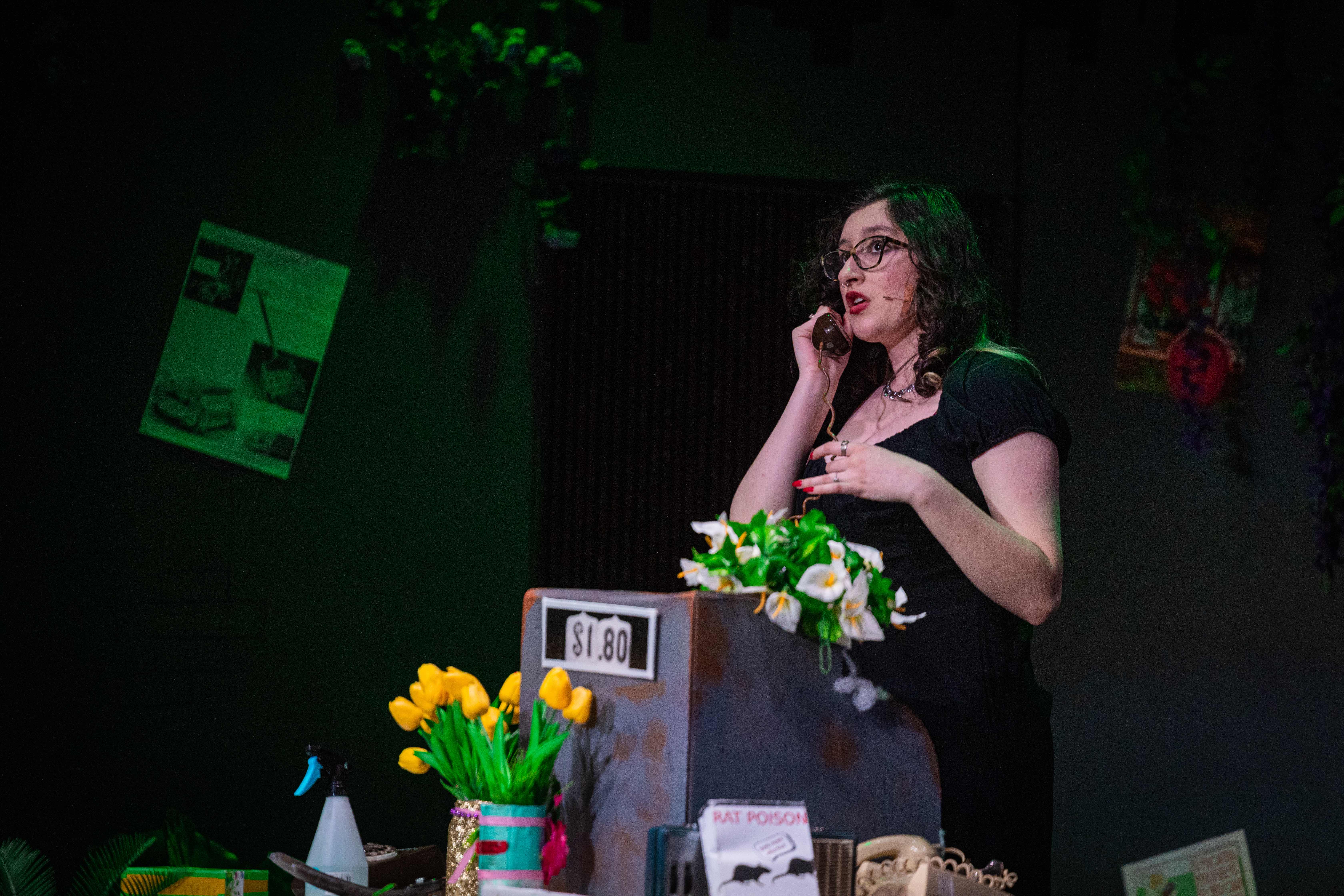 A young woman stands on stage holding a phone to her ear. She stands among a flower shop