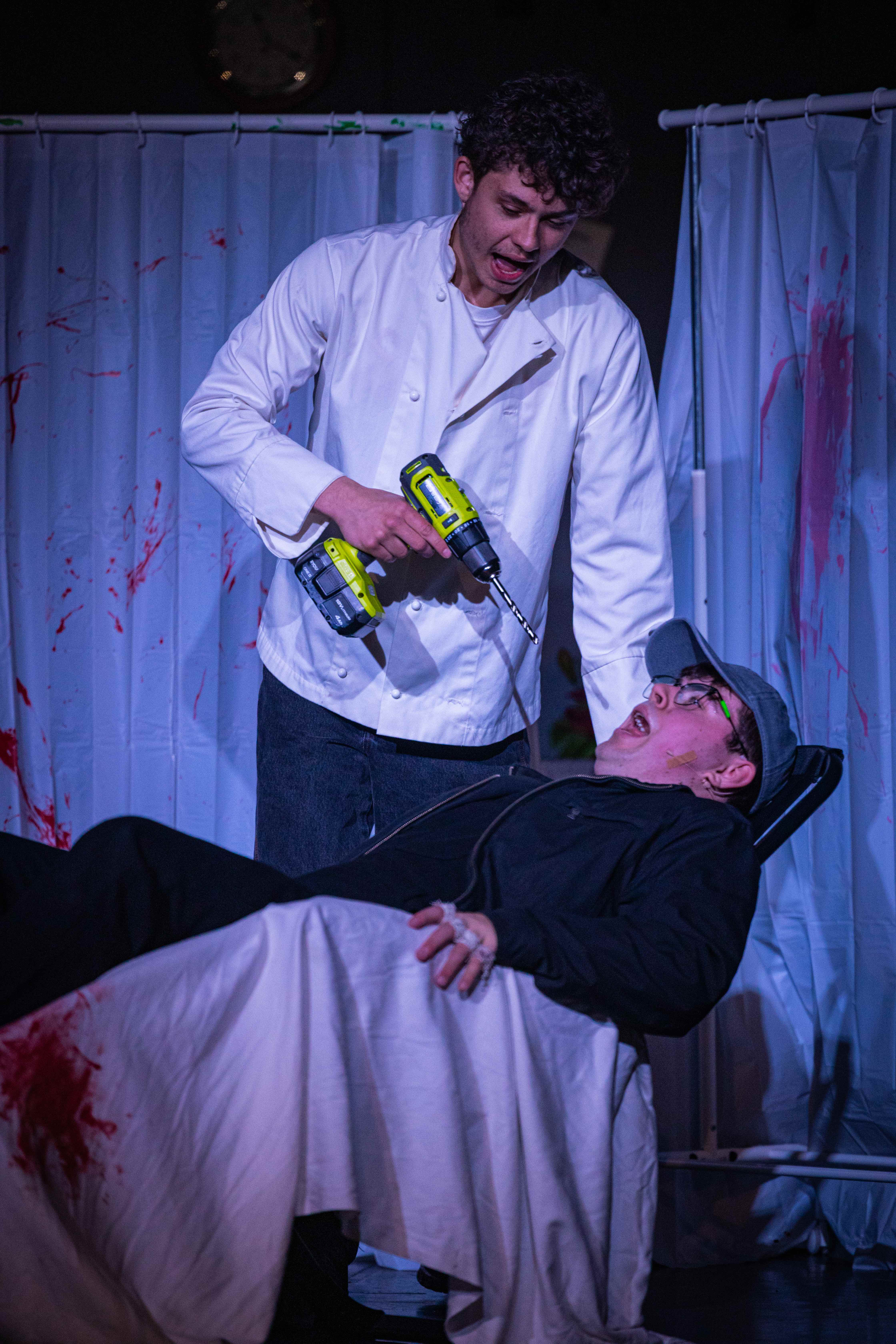 Two young men stand on stage acting. One of the men is dressed as a dentist and points a drill at the other man who lies in a dentist's chair