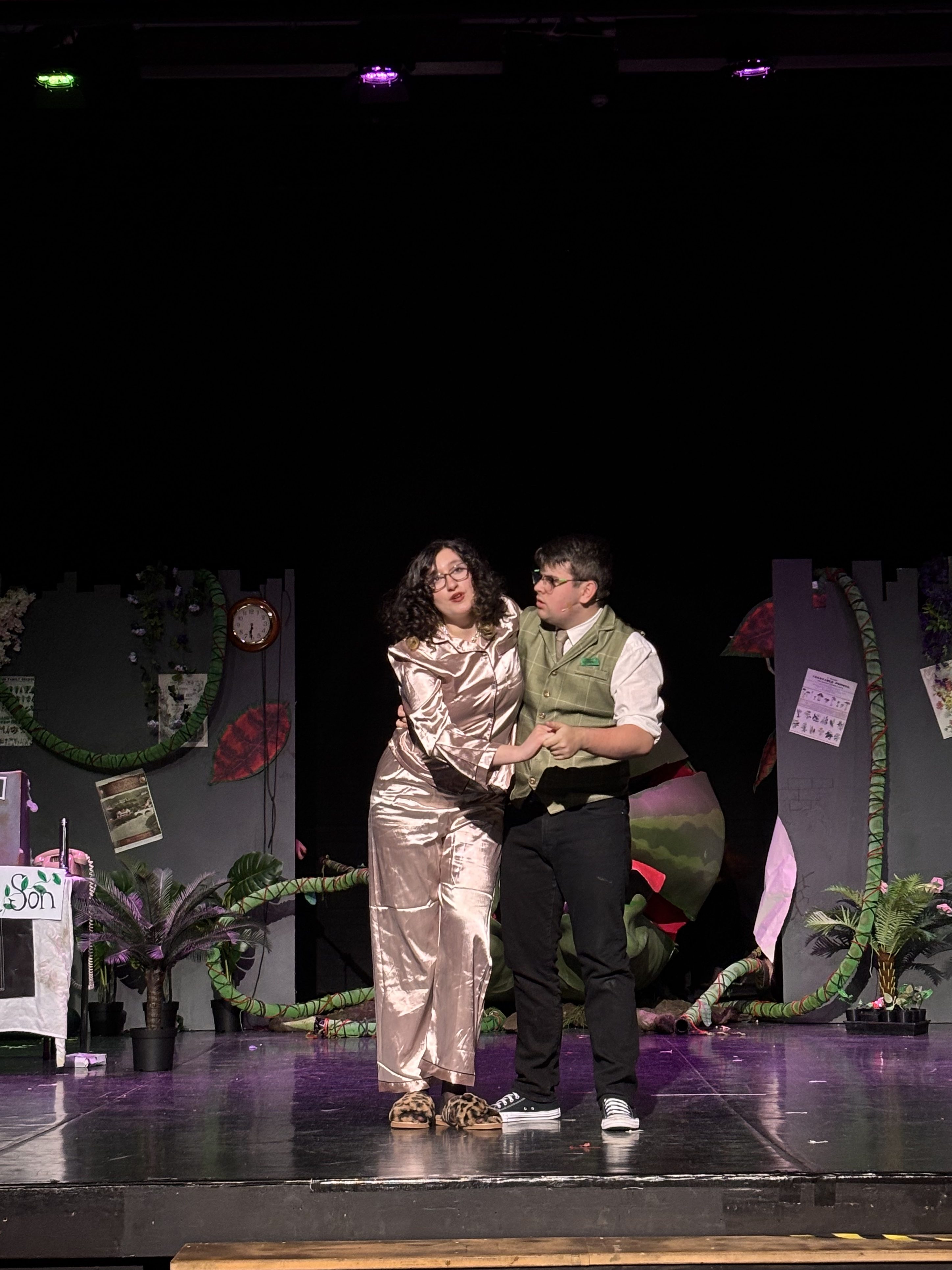 A young woman wearing silk pyjamas is held up by a young man. They stand in front of a fake plant shop