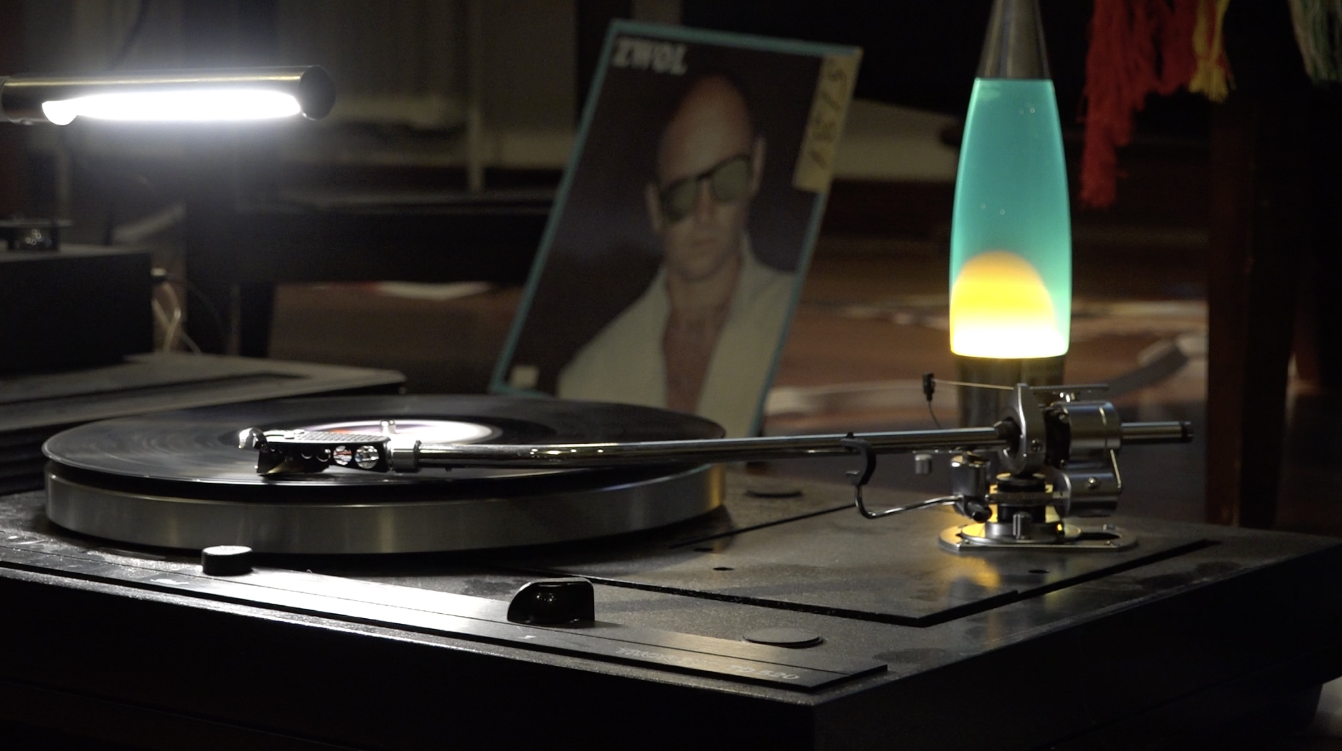 A black vinyl is played on a black record player. In the background there is a blue and yellow lava lamp and a record cover.