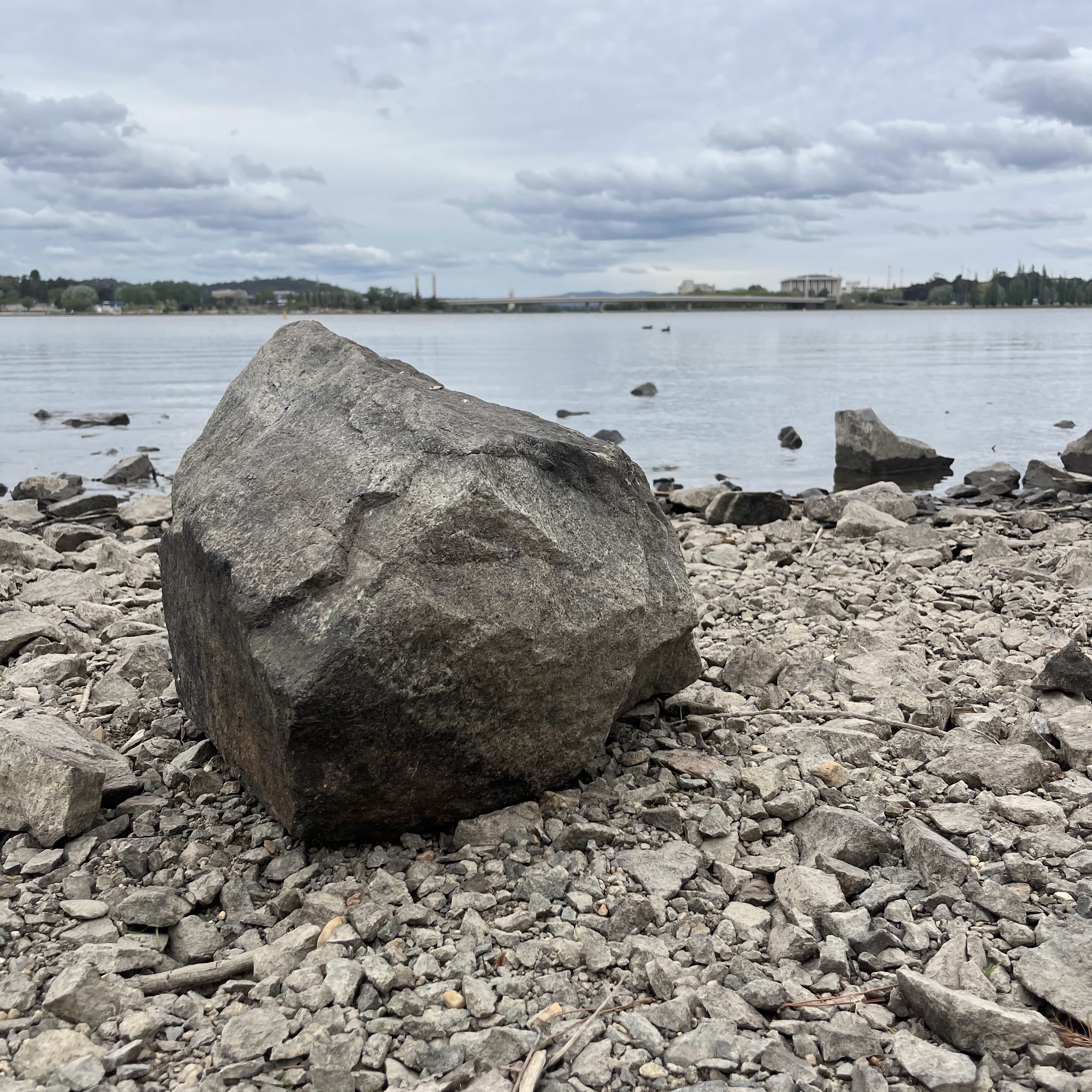 Large boulder sits ontop of pebbles by the side of a lake.