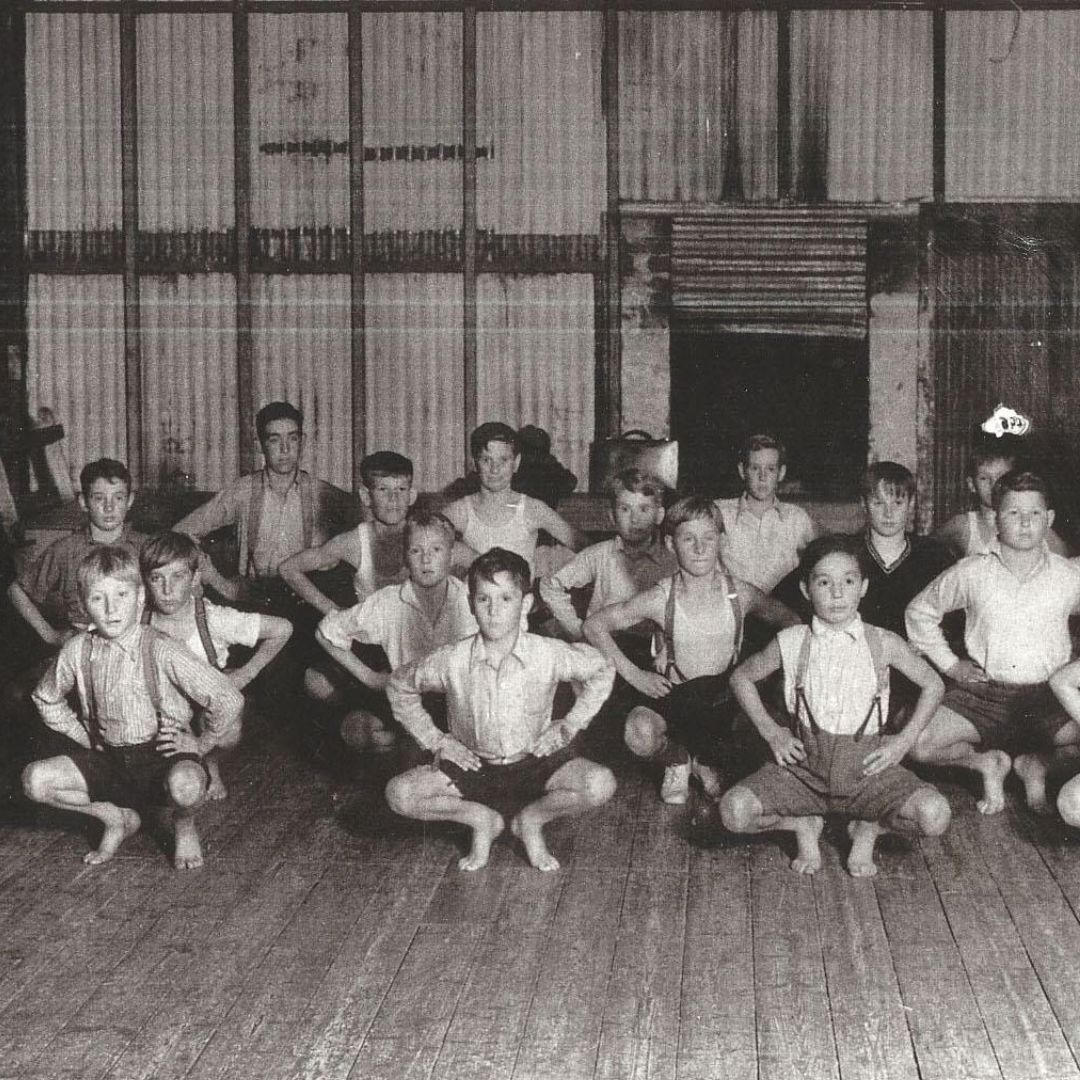 A group of young boys in shorts, shirts and braces squat inside a community hall with corrugated walls.
