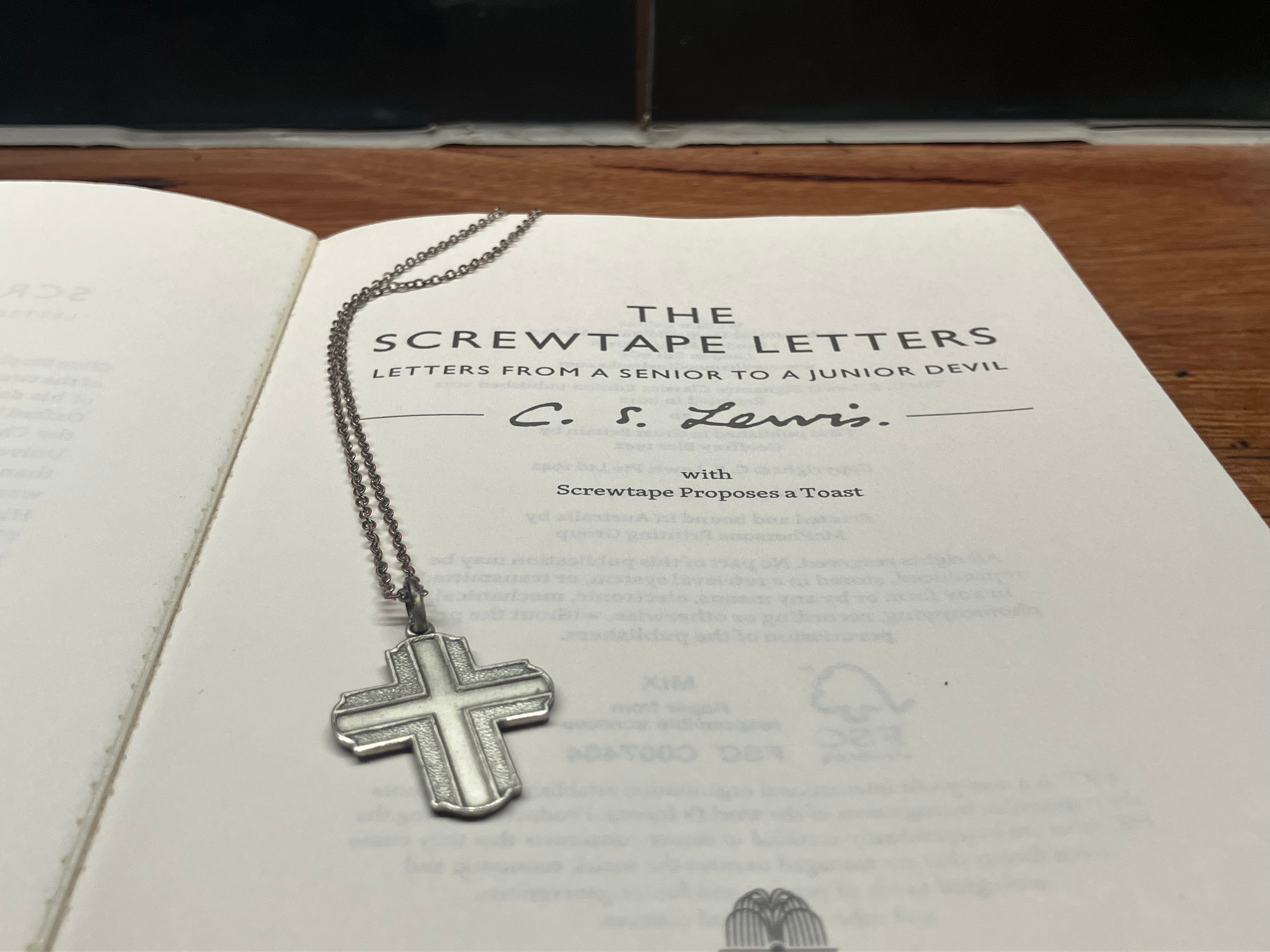 The open title page of The Screwtape Letters sitting on a wooden counter with turquoise tiling behind. There is a large silver crucifix lying down the left side of the page.