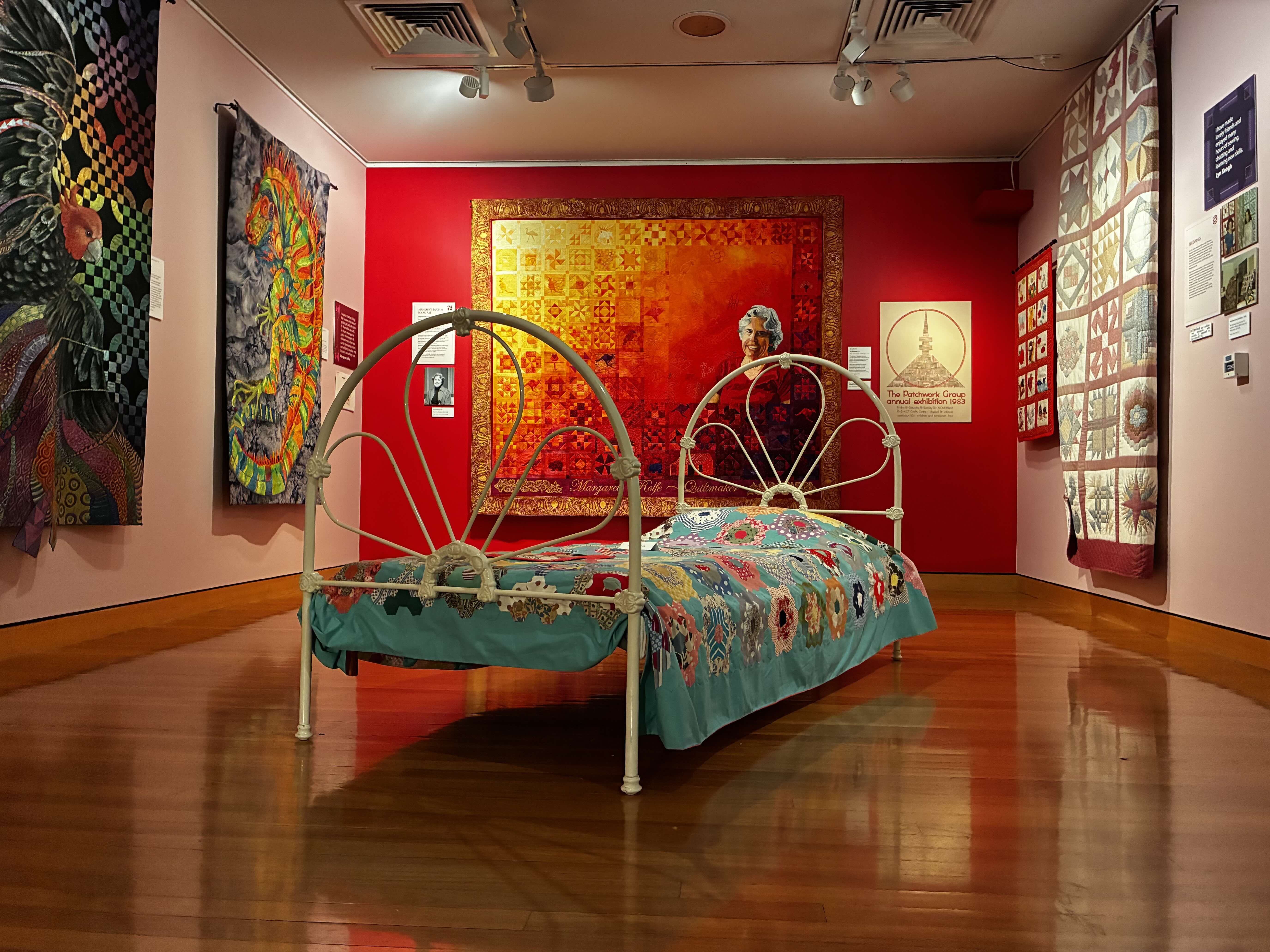 A white metal bed with blue quilt covered in multicoloured hexagons. The bed sits in the middle of the exhibition space.
