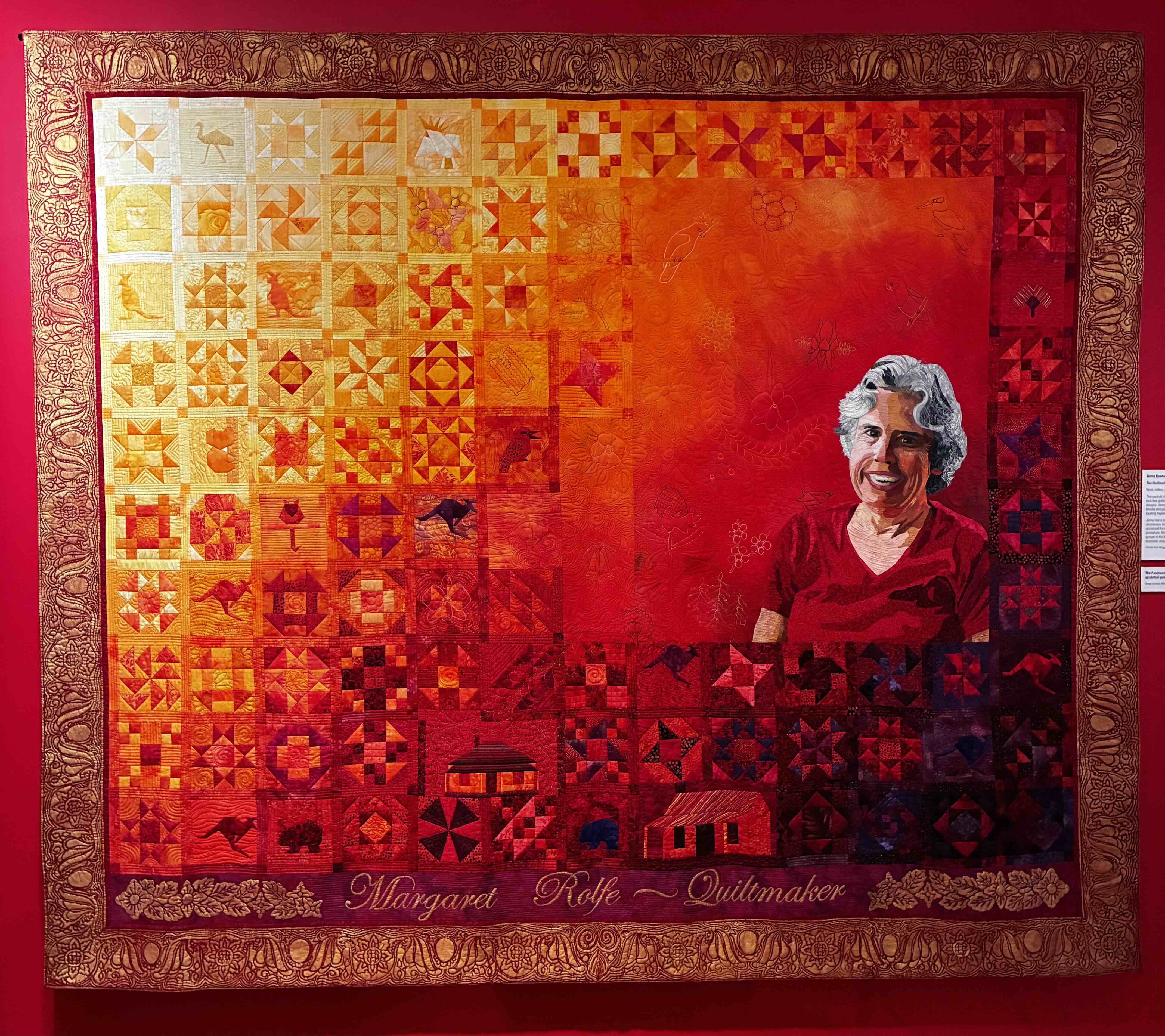A large quilt made of squares that transition from yellow and orange to red and purple. Each square has a geometric design. There is a colour portrait of Margret Rolfe stitched into the quilt. The edges of the quilt are painted gold.