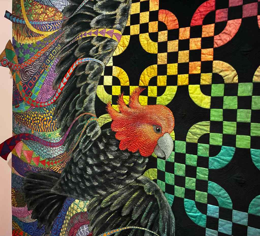 A portion of a large quilt with black and white Gang Gang cockatoo. The cockatoo has multicoloured ribbons in its wing. It is flying on a black and rainbow check background.