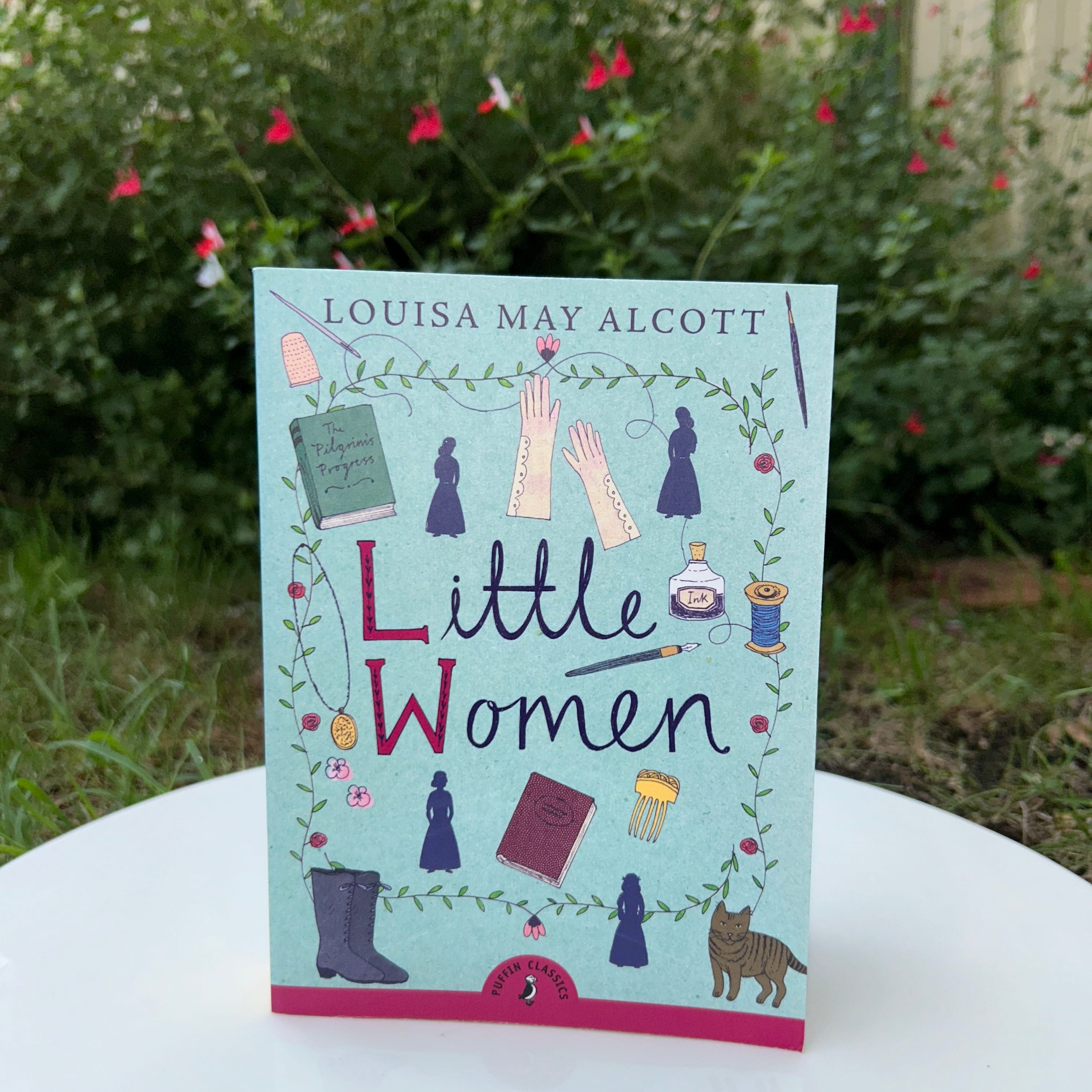 The Little Women book sitting in front of a flower bush.