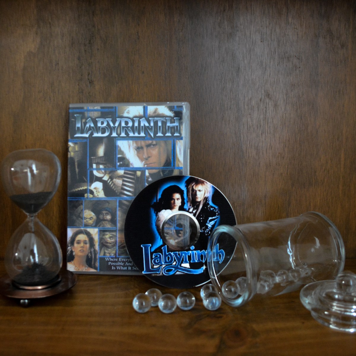A DVD copy of 'Labyrinth' sitting on a shelf adorned with an hourglass and a spilt jar of clear glass marbles.
